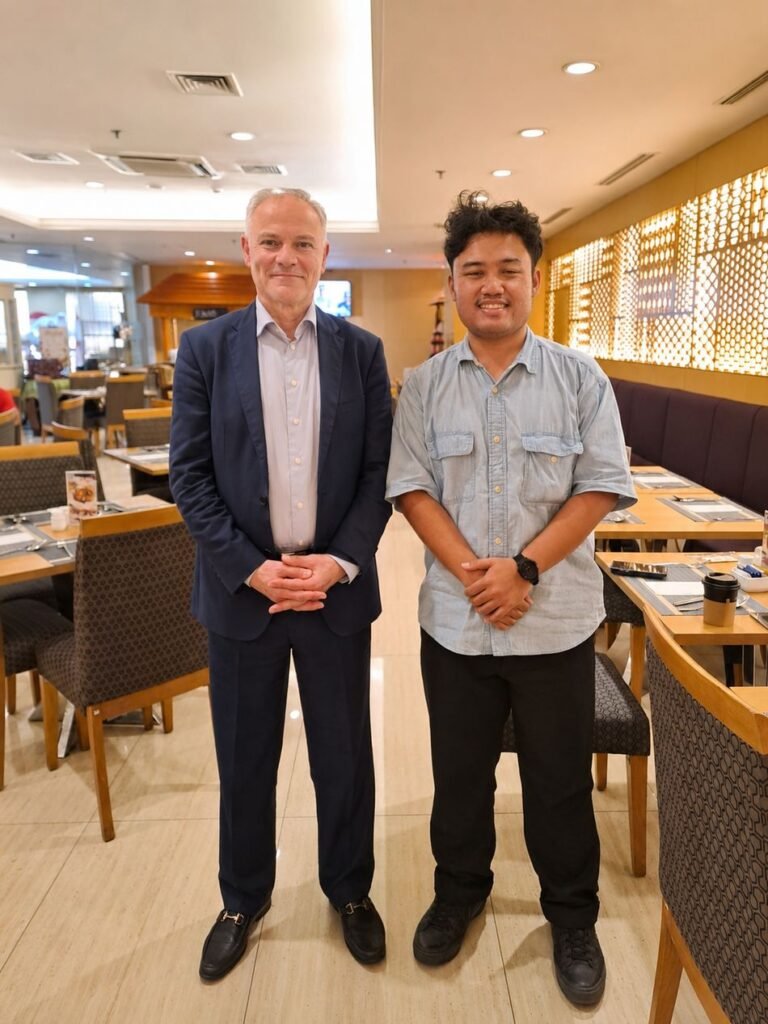 Two men stand side by side for a photograph in a restaurant, one in a navy suit and the other in a light denim shirt.